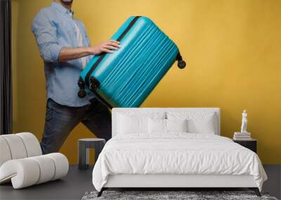 Full body photo of a funny young happy man in sunglasses carrying his blue suitcase jumping and having fun on a yellow background. Summer holidays, vacation trip and travel concept. Wall mural