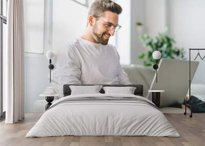 A Young attractive guy is browsing at his laptop, sitting at home on the cozy beige sofa at home, wearing casual outfit Wall mural