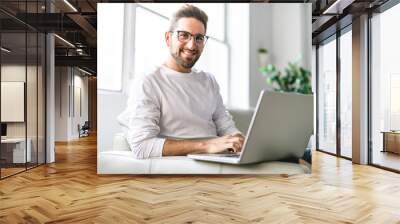 A Young attractive guy is browsing at his laptop, sitting at home on the cozy beige sofa at home, wearing casual outfit Wall mural