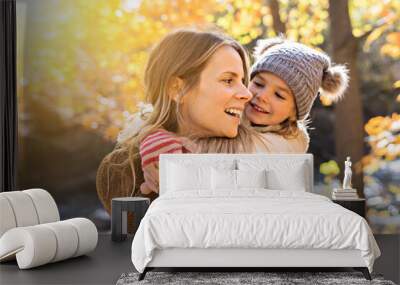 Little girl and her mother playing in the autumn park with child on back Wall mural