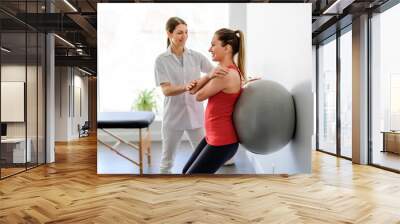 Physiotherapist helping woman do wall squats with fit ball. Young female patient doing back exercise using fitball in physio room of modern clinic Wall mural