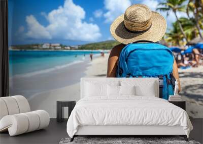 A woman with a blue backpack and a straw hat walks along a white sandy beach towards the turquoise ocean. Wall mural