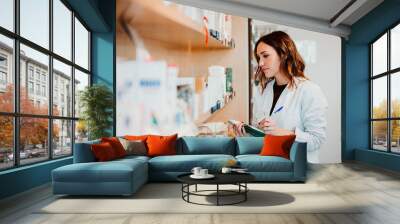 .Young female pharmacist working in her large pharmacy. Placing medications, taking inventory with her green notebook. Lifestyle Wall mural