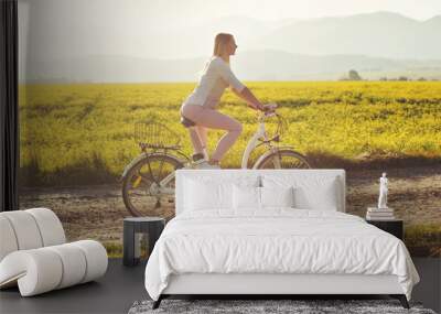 Young woman rides electric bicycle on dusty country road, view from side, sun backlight field with yellow flowers background Wall mural