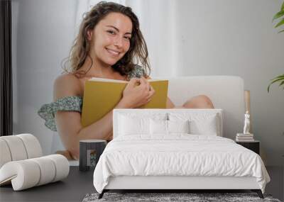 Young attractive woman sitting in her living room armchair reading her favorite book. Reading is a way of learning new things. Reading is a very entertaining and exciting hobby. Relaxed young woman. Wall mural