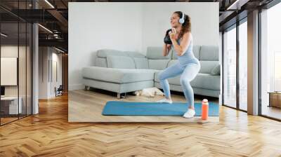 Young woman exercising at home with her pet. Listening to music with her headphones. Motivation to achieve results. Practicing sports is good for the body and mind. Healthy body, healthy mind Wall mural