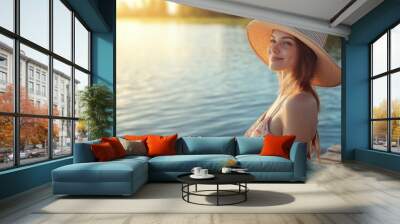 A pleased woman in a classic sunhat and a summer dress, standing on a wooden pier, gazing at the calm water in the sunlight Wall mural