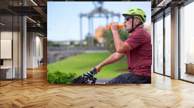 Active senior man wearing cycling helmet drinking an orange juice from a bottle riding the electric bicycles in the city. Authentic retirement living and healthy lifestyle concept. Wall mural