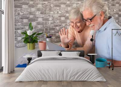 Attractive elderly couple smile while video chat by laptop with family or friends, two attractive elderly people using computer at home drinking a cup of coffee or tea Wall mural