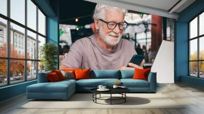 Attractive senior man sitting in a cafe smiling while using mobile phone. Cheerful senior male, using modern technology expressing happiness and contentment in a relaxed atmosphere Wall mural