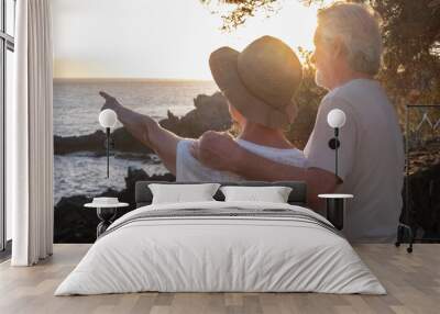 Back view of two happy and romantic seniors or pensioners embraced at the sea at sunset light looking at horizon - old couple outdoors enjoying vacations together Wall mural