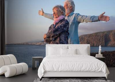 Carefree senior couple standing on the rocks at sea looking at the horizon over water at sunset light. Relaxed lifestyle for a caucasian couple of retirees Wall mural