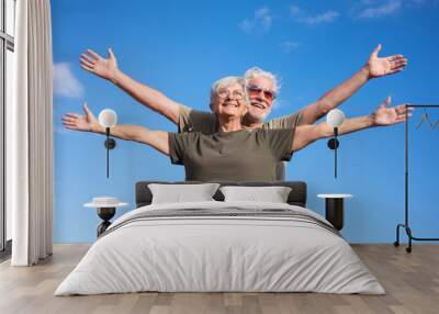 Caucasian couple of two happy senior people enjoying outdoors and nature, standing with open arms feeling freedom. Smiling white-haired man and woman wearing sunglasses, blue sky, copy space Wall mural