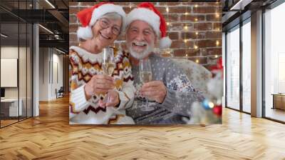 Cheerful senior adult couple at home enjoying christmas together in december wearing santa hats. Man and woman toasting with sparkling wine glasses looking at camera in holiday leisure. Xmas tree Wall mural