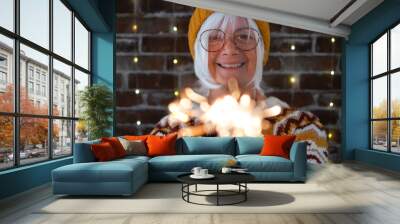 Handsome senior caucasian woman with eyeglasses and winter sweater holding sparkler light, elderly happy lady celebrate Christmas and new year sparkling in the night Wall mural