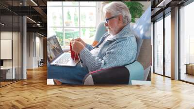 Handsome senior man learning how to knitting work with woolen yarn at home watching an online class tutorial on laptop computer. Concept of female hobbies for male people. Modern lifestyle people Wall mural