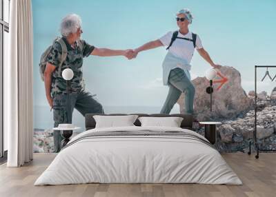 Happy active senior couple on outdoor hike in mountains helping each other up enjoying freedom and healthy lifestyle. Vacation or retirement. Horizon over water background Wall mural