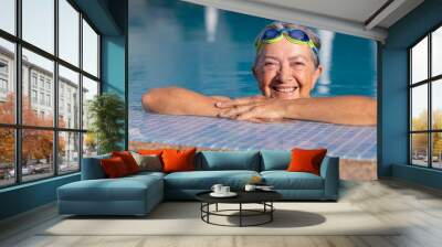 Happy active senior woman doing sport in the outdoor swimming pool looks at camera smiling. Elderly woman and healthy lifestyle in retirement or vacation Wall mural