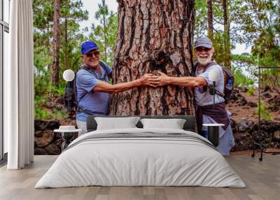 Happy couple of senior friends in mountain excursion looking at camera laughing while embracing a large tree trunk, two retired people enjoying healthy lifestyle in nature Wall mural