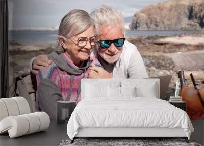 Happy senior couple browsing together on laptop enjoying sunny day outdoor at the beach sitting at bar wooden table with orange cocktail. Elderly people in vacation or retirement at sea Wall mural