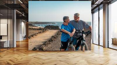 Happy senior couple of cyclists running bicycles in footpath at sea looking each other. Elderly man and woman enjoying freedom and healthy lifestyle Wall mural