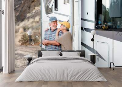 Happy senior couple on a leisure trip standing outside a camper van tenderly embracing. Handsome retired caucasian couple enjoying freedom and alternative life in camper van Wall mural