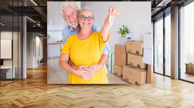 Happy smiling couple of senior people white hair hugging  holding the keys of the new empty apartment with moving boxes on the floor - concept of active elderly people and new beginning like retired Wall mural