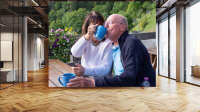 Joyful senior couple sitting at a wooden cafe table outdoors in the mountain enjoying a cup of hot tea. Elderly traveler couple spending free time together Wall mural