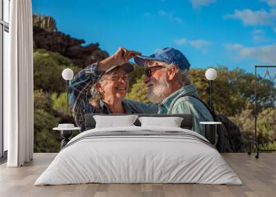 Lovely happy senior couple hugging in mountain trekking enjoying nature, freedom and healthy lifestyle. Smiling old retirees in hat and casual clothes among green bushes and blue sky Wall mural