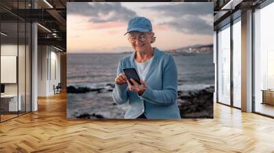 Portrait of beautiful senior woman in blue cap sitting outdoors at beach in sunset light using mobile phone. Smiling senior woman texting on mobile phone Wall mural