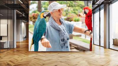 Portrait of mature elderly woman in hat and sunglasses holding on her arms two domestic ara parrots in the park Wall mural