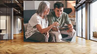 Senior woman and young grandson watch laptop together at home. Young boy teaches computer technology to his elderly grandmother how to surf, buy on internet. Elderly woman learns how to use computer. Wall mural