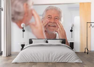 Skin care. Handsome senior man applying cream at his face and looking at himself with smile while standing in front of the mirror Wall mural