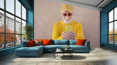 Smiling caucasian senior man in yellow with sunglasses and hat holding a smart phone looking at funny videos Wall mural