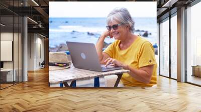 Smiling elderly woman sitting outdoors at cafe table using laptop. Active blogger nomadic worker Wall mural