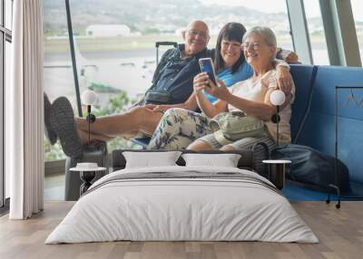 Smiling group of senior friends sharing tech and social on mobile phone sitting in airport departure area waiting for boarding. Travel and tourism concept, retiree lifestyle Wall mural