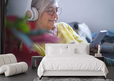 Smiling relaxed senior woman wearing headphones sitting on sofa at home using smart phone looking at social news Wall mural