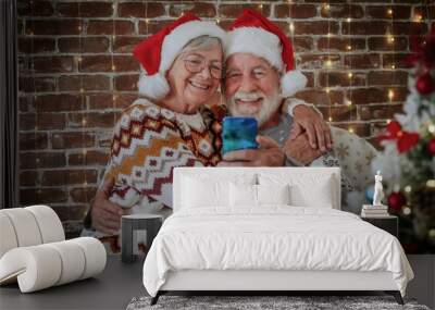 Smiling senior adult couple at home hugging enjoying christmas together at home wearing santa hats. Man and woman video chatting with distant family or friends by phone. Xmas tree Wall mural