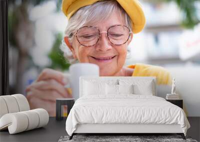 Smiling senior woman having relaxed moment with an espresso coffee cup at outdoor cafe looking at mobile phone. Attractive caucasian elderly lady in yellow holding white coffee cup Wall mural