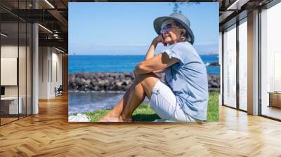 Smiling senior woman in straw hat sitting barefoot in meadow face the sea listening to music, relaxed senior lady enjoying free time in summer vacation or retirement Wall mural