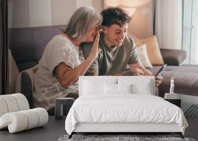 Video call concept. Happy senior retired woman sitting on sofa with young nephew waving hands video calling by mobile phone using technology for online webcam connection with distant family or friends Wall mural