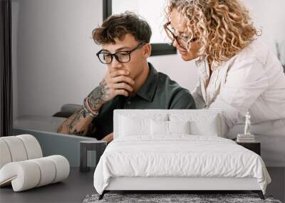 Young smiling boy studies at home with the support of a personal tutor using laptop. Blonde teacher follows the instruction of a handsome boy. Concept of modern education with the use of technology Wall mural