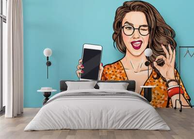 Winking woman in glasses showing smart pone and OK sign. Pop art girl holding phone. Digital advertisement female model showing the message or new app on cellphone. Wall mural