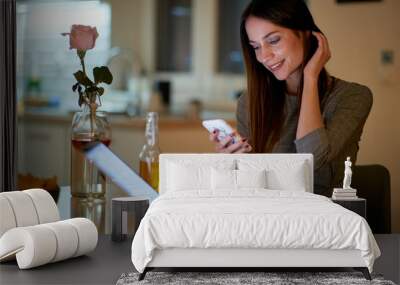 A young beautiful girl with a drink sitting at the table in an apartment and chatting on a smartphone. Leisure, date, meeting Wall mural