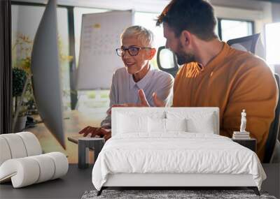 A young businessman and elderly female colleague have a conversation about a computer screen content watching together at the desk at workplace. Business, office, job Wall mural