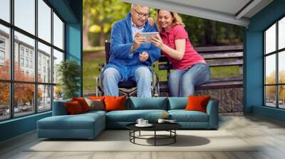 daughter with her disabled father in wheelchair using a digital tablet, talking and smiling. Wall mural