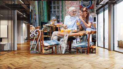 elderly woman showing part in opened book to young adult brunette female drinking orange juice on a straw, sitting in outdoor cafe Wall mural