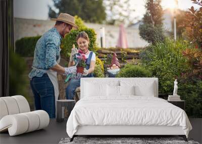 Father and young girl child assisting with re planting a flower from a flower pot, sitting by the flower garden by the house, feeling joyful. Wall mural