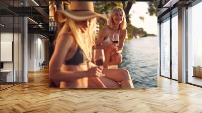 Girlfriends by water, having small talk, holding drinks. Young beautiful women sitting on wooden jetty. Vacation, summer, fun, holiday, lifestyle concept. Wall mural