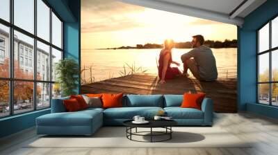 young couple sitting on wooden platform by the lake, looking each other, talking at sunset Wall mural